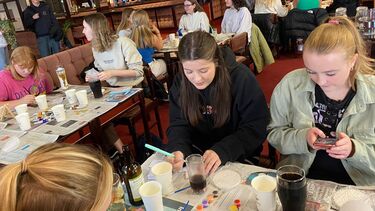 Two people shown sat at a table painting tiles