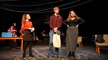 three students on a stage, one on the right speaking with hands on hips