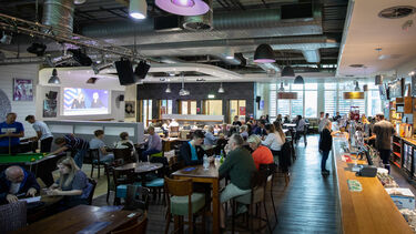 A room full of tables and chairs where people are sat eating and drinking, there is a bar to the right hand side