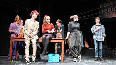 Informal scene from a stage play in which some actors are sitting on a table and they look in the direction of the speaker to the right.