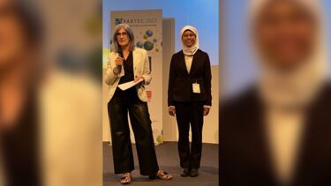 Yang Sarah Mohamad smiling by a poster for PARTEC with another person