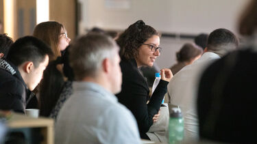 A woman with curly brown hair and dark rimmed glasses is sitting in a lecture theatre audience listening attentively