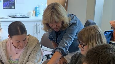 Blod woman leaning pointing at something on a table while two students look and smile