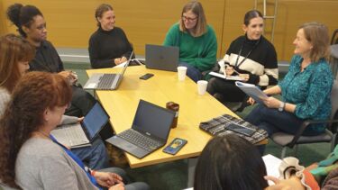 A group of people sat around a table in discussion, with laptops and notepads. 