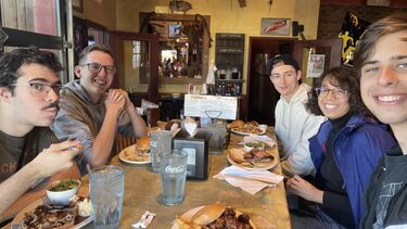 Five students sit around a table with plates of barbecue food next to them. They are all smiling at the camera.