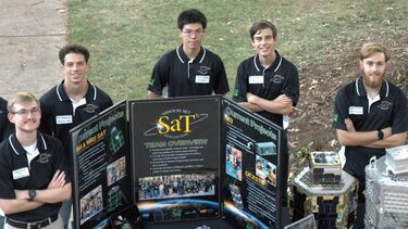 A group of five male students stand outside around a small table that is promoting university opportunities to prospective students. They are all smiling towards the camera.
