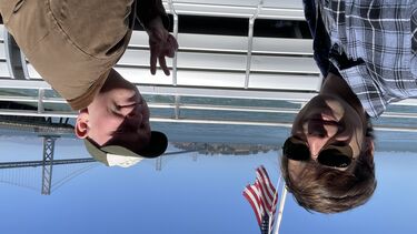 Two male students sit on a ferry with the American flag behind them and a view of the Golden Gate Bridge in the distance. They both smile at the camera.
