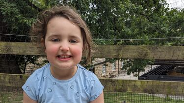 Zoe is a little girl with brown hair, she sits in a park and smiles