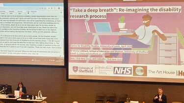 Two large screens sit behind Kirsty Liddiard who is delivering a keynote lecture. The back of people's heads in the audience are visible. 