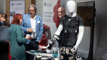 People talking at an exhibition stand. In the foreground there is a white mannequin dressed in black clothing and a variety of wearable sensors