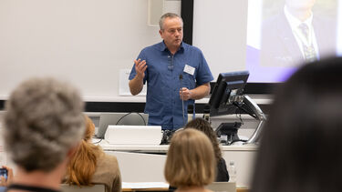  A man wearing a blue shirt is stood in front of a lectern talking to an audience.