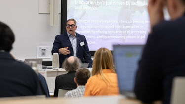 A man in a dark suit and dark frame glasses is talking in front of a lecture with a slideshow behind him to a lecture theatre full of people