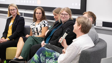 A group of people sat on a sofa looking towards a woman speaking into a microphone