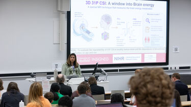 A woman standing in front of a lecture speaking to a lecture theatre full of people with a scientific slideshow on the wall behind her