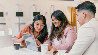 Three students sat at a table looking at a shared laptop screen