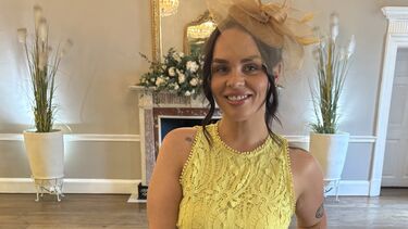 Becky wears a yellow dress and a fascinator. Her hair is dark brown/black and styled for a formal event.