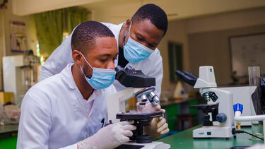 Two people in a lab looking through a microscope, wearing white coats and masks.