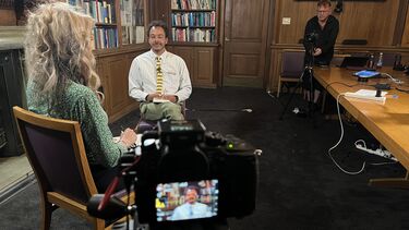 Studio set up with two poepl sat opposite each other in an interview setting and the filmer in the background. The setting is traditional with a lot of dark wood. 