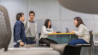 A group of four students stand and sit around a table and talk with one another