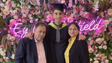 Student Vayam Lahoti wearing graduation cap and gown, with a man and a woman, stood in front of fake flowers and a neon sign