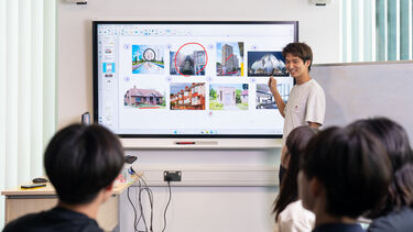 A tutor stands at the front of a classroom with an interactive whiteboard behind them and smiles at the students.