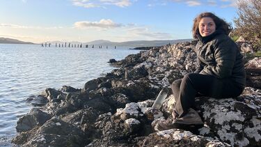 Student Alicia Rowe sits on rocky shoreline beside the sea, smiling at the camera. She wears a black jacket and trousers, with a blue sky and clouds above the water in the background.