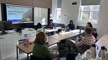 Five people sat around some tables having a discussion in front of a presentation screen