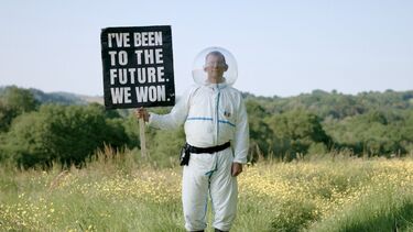 Rob Hopkins standing in a feild wearing a spacesuit and holding a sign that reads "I've been to the future. We won."