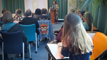 ebecca sits with a sketchpad at the back of an audience, facing a women giving a speech.