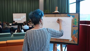 View from behind of a woman with blue hair drawing on an easel, stood behind the symposium audience.