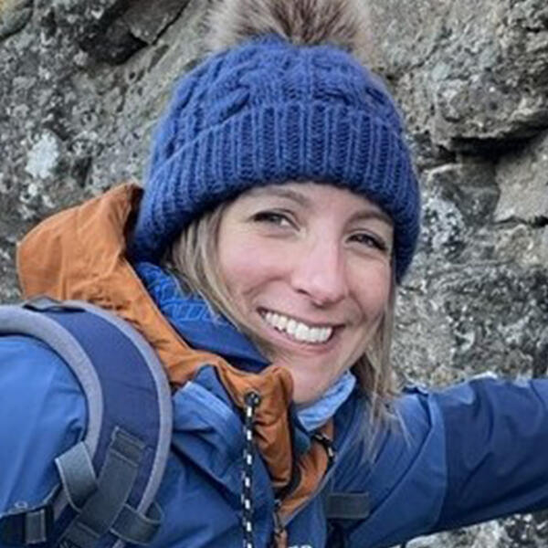 Profile picture of Helen wears a blue bobble hat and blue jacket, and she stands in front of a stone wall.
