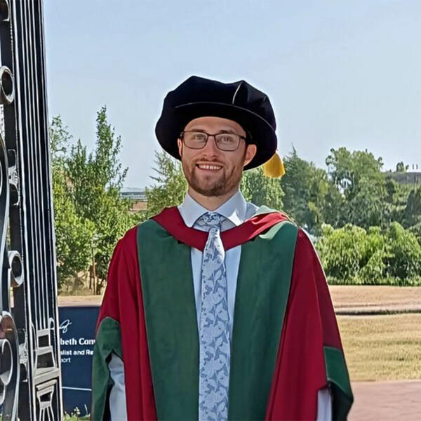 Profile picture of Liam Hughes, a white male, stands wearing graduation gown, hood and cap