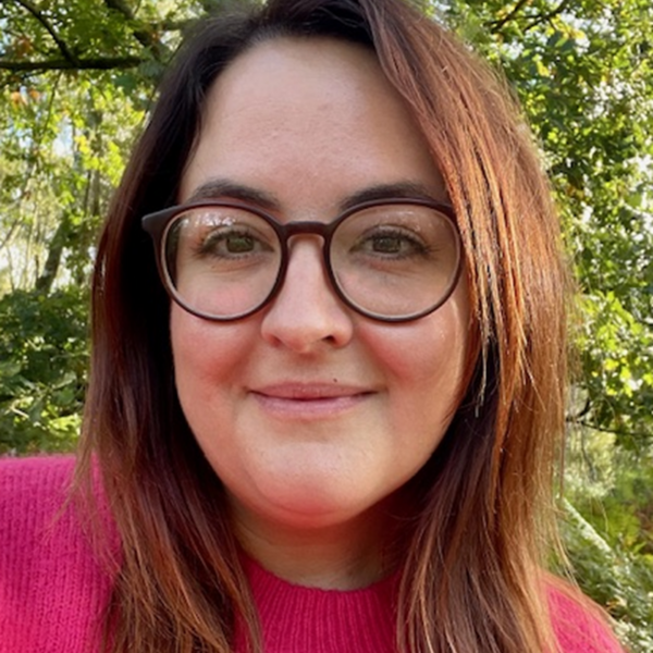 Profile picture of Emma faces the camera in front of a tree, wearing glasses and a pink jumper