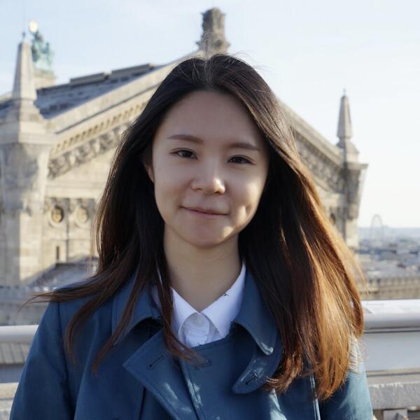 Profile picture of Nan (Elva) Peng smiles at the camera, with buildings in the background
