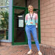 Amy Harding wears a pastel ombre jumper and skinny blue jeans. She has blonde hair. She stands in front of a glass panelled building.