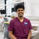 Randeep has dark curly hair, and wears burgundy dental scrubs. He sits on a chair next to a row on phantom head simulators.