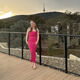 A female student stands outside on a wooden pathway, with a railing behind her and smiles towards the camera. She is wearing a pin occasion dress and behind her is a backdrop of green hills and trees.
