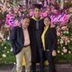 Student Vayam Lahoti wearing graduation cap and gown, with a man and a woman, stood in front of fake flowers and a neon sign
