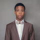 A male university student stands and looks towards the camera. There is a grey background.