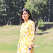 Shriya Bajaj standing on a green field in a yellow dress with trees in the background