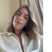 Kate looks down at the camera, wearing a white shirt, with a curtain in the background