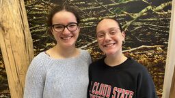 Two young women smiling at the camera standing in front of a nature poster
