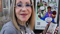Margaret Colling, a retired Librarian, holding her phone up at a bus stop with the Let's Talk AI campaign webtoon in the background.