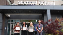 Three students smiling and holding certificates with lecturer outside the medical school.
