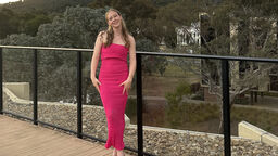 A female student stands outside on a wooden pathway, with a railing behind her and smiles towards the camera. She is wearing a pin occasion dress and behind her is a backdrop of green hills and trees.